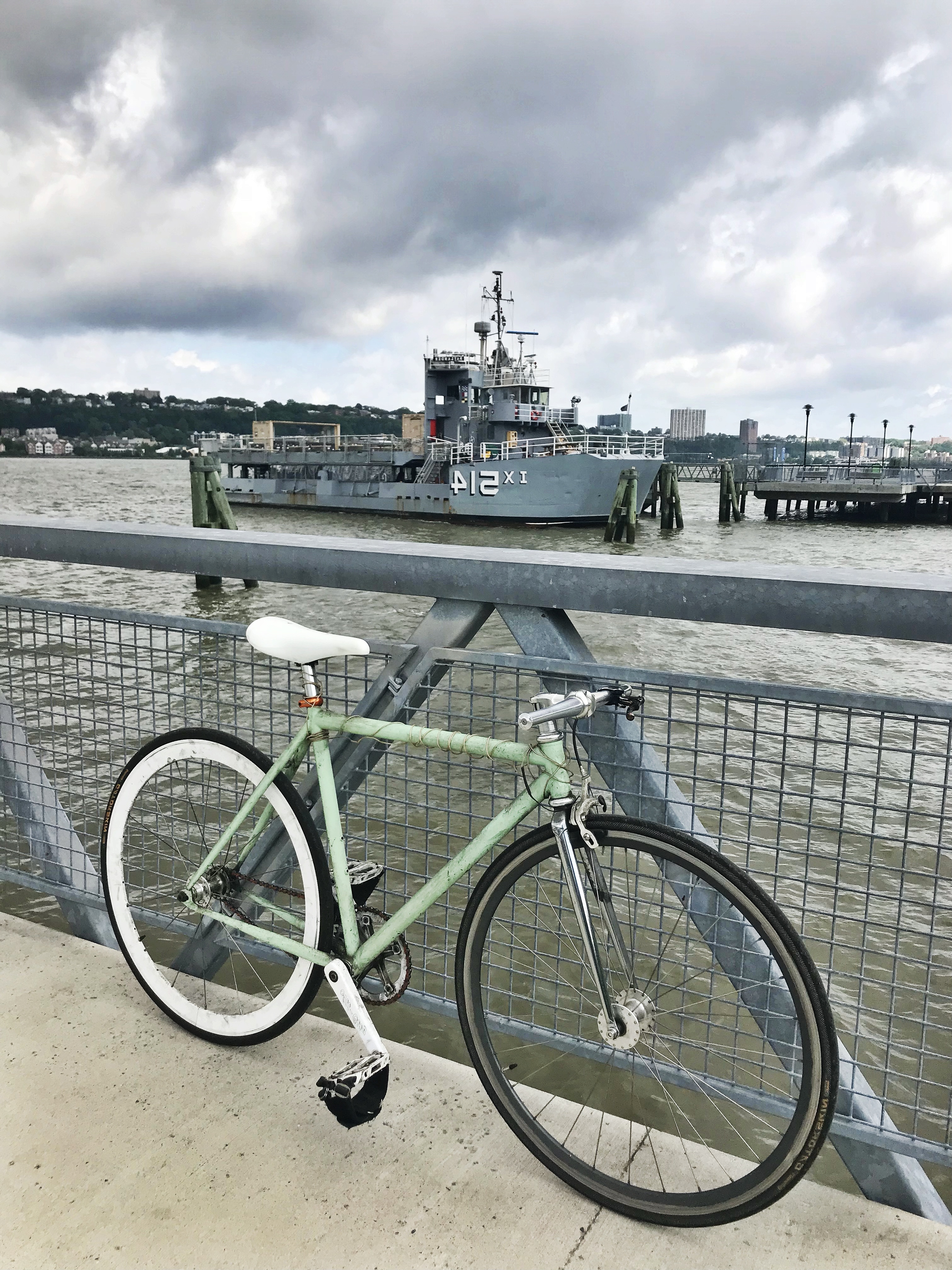 Mint green single-speed bike with hipster vibes leaning against a metal barrier, set against the Hudson River on a gloomy day