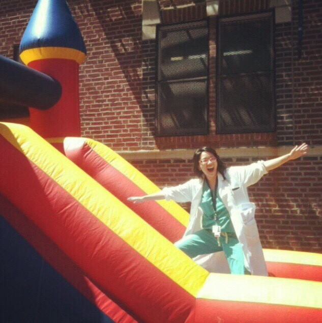 Young Korean American woman with glasses wearing mint green scrubs and a white coat, arms outstretched, standing on a giant inflatable castle