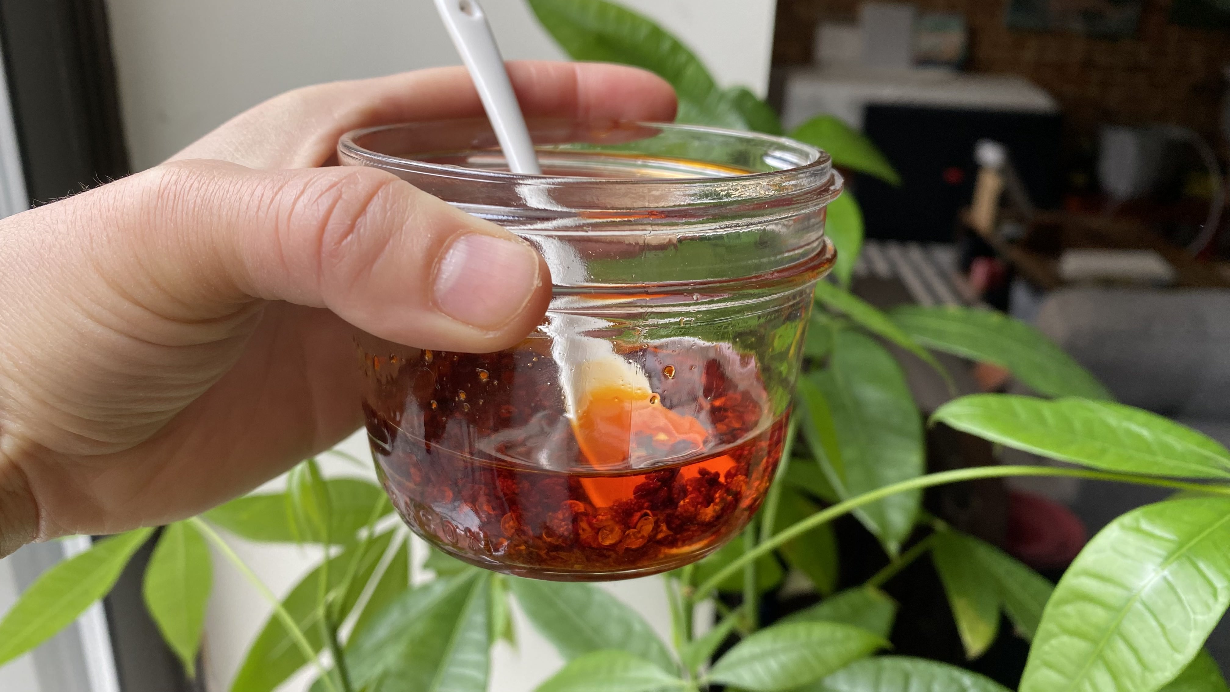 A short glass jar about one-thirds full with orangey red chili oil, with Sichuan peppercorns and gochugaru throughout