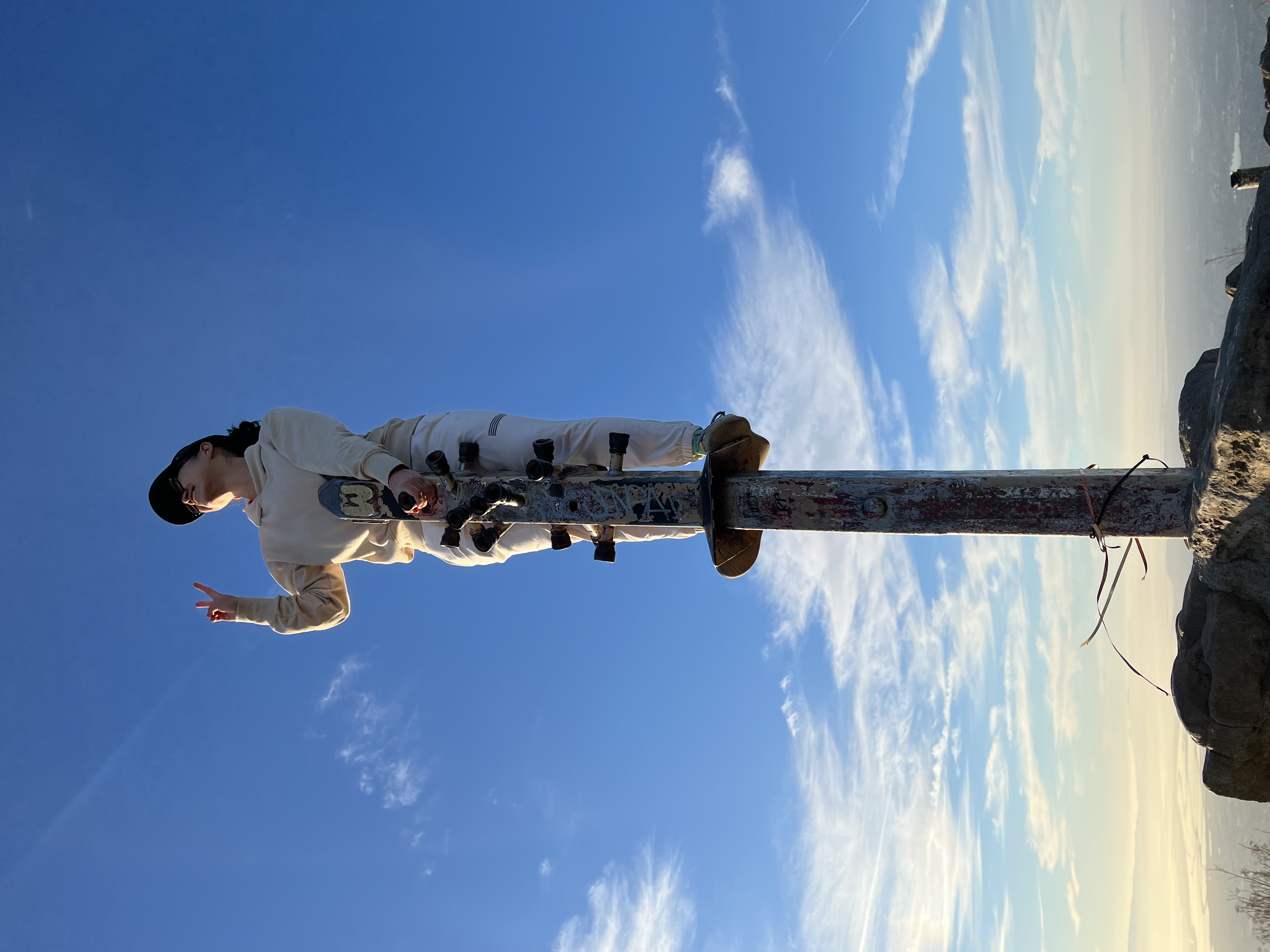 Person standing on a pole with horizontal protrusions acting as footrests similar to a pogo stick, atop a hilly peak. The background is blue sky with a few wispy clouds.