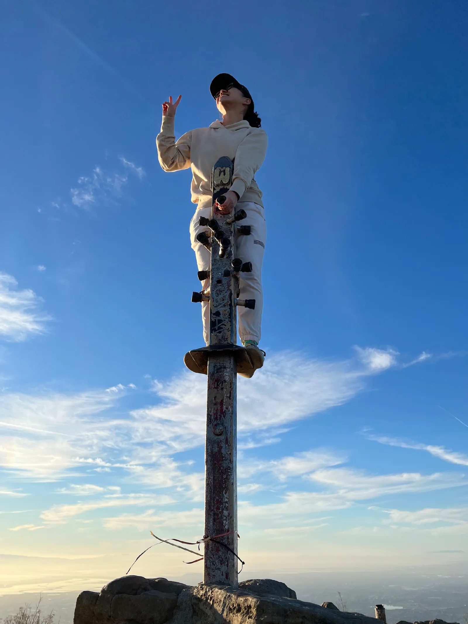 Person standing on a pole with horizontal protrusions acting as footrests similar to a pogo stick, atop a hilly peak. The background is blue sky with a few wispy clouds.