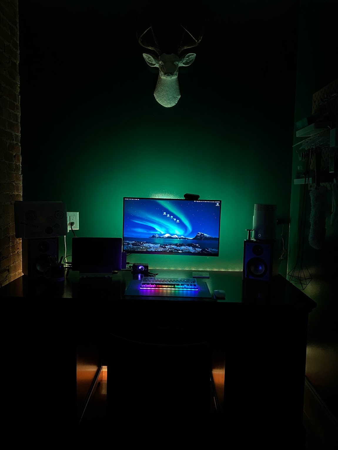 A dark and liminal room with a deep green wall on which hangs a fake white deer head with gold antlers; on the desk is a computer setup with a glowing, backlit monitor and mechanical keyboard