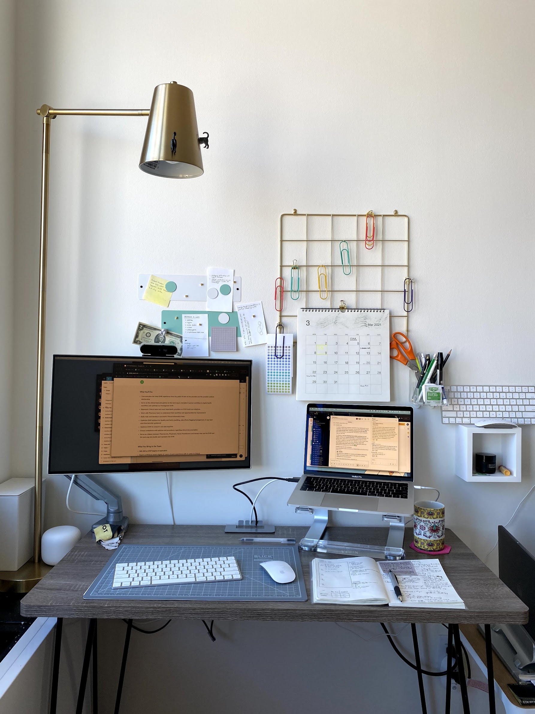 An office setup in the naturally-lit corner of a bright room with white wall. An open Macbook Pro and computer monitor sit on the writing desk, with various other stationery items scattered about