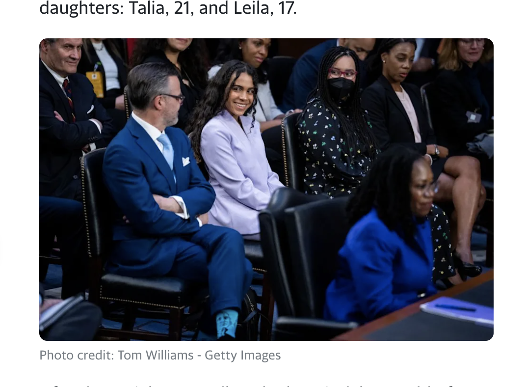 Screenshot of a news image of a courtroom photo showing Hon. Ketanji Brown Jackson's husband sitting with one leg crossed over the other. He is wearing a suit and a bright blue sock is visible under his pant leg.