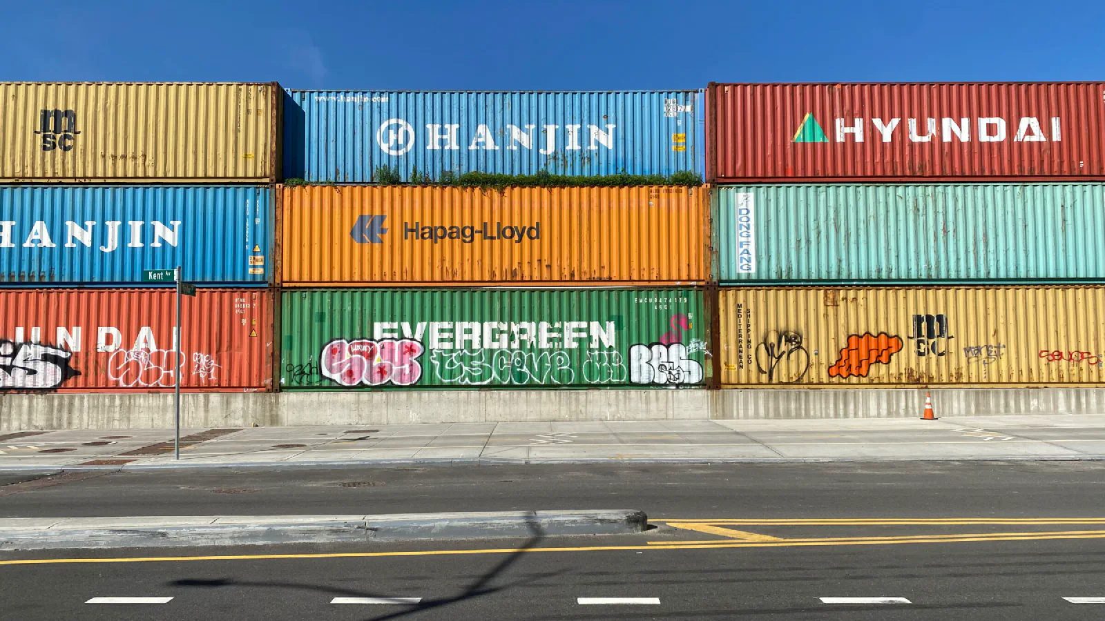 Head-on view of a several colorful shipping containers, stacked 3-high, along an empty roadside against the blue sky